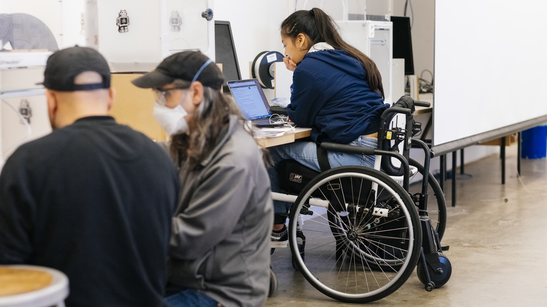 a woman in a wheelchair using a desktop computer while two men share a computer